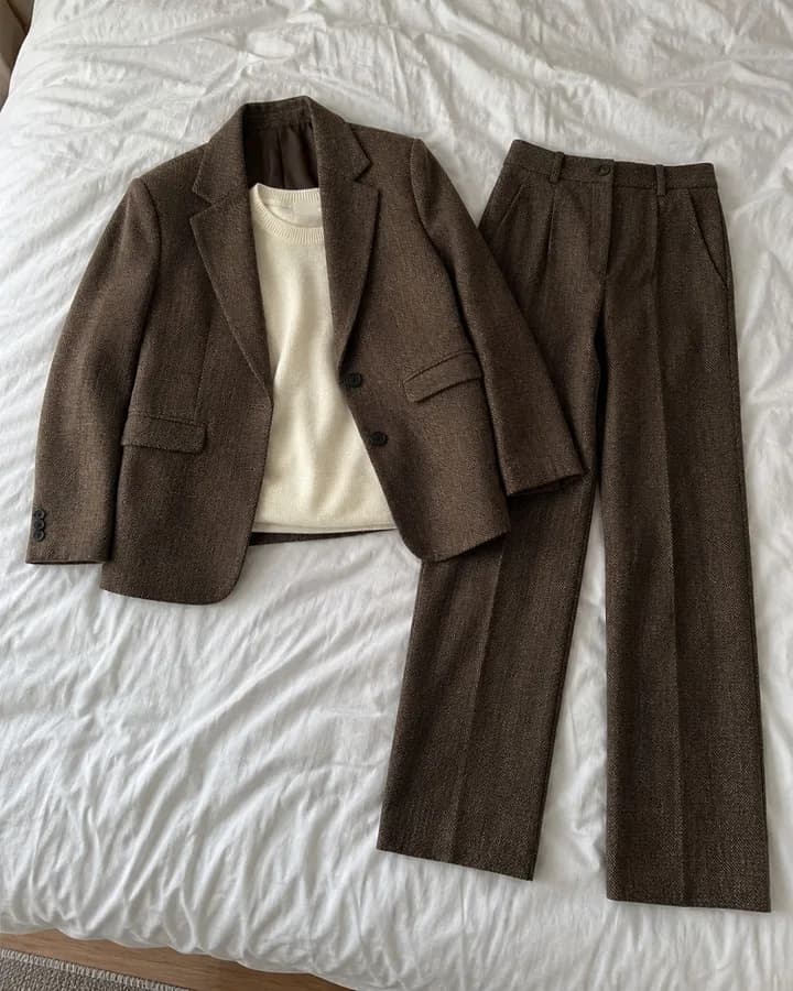 Flat lay garment photo: a brown herringbone blazer, cream knit top and matching trousers laid out on a white bed sheet