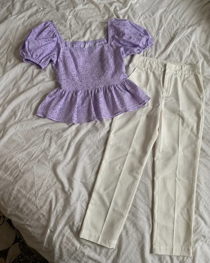 Flat lay garment photo: a lilac eyelet peplum top and white trousers laid out together on a bed